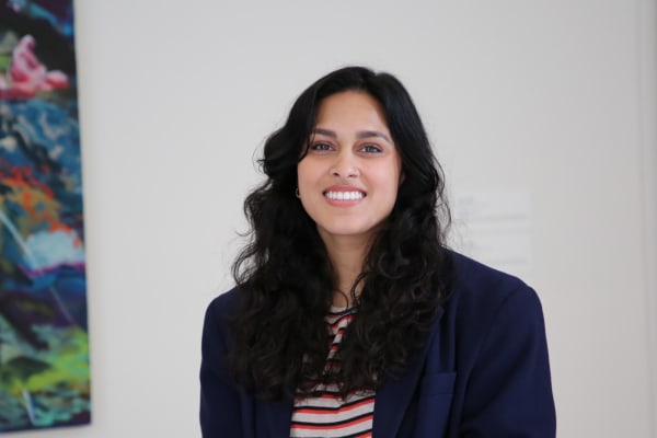 A portrait of a smiling woman with long, dark curly hair, wearing a striped shirt layered under a dark blazer. She has a nose ring and appears relaxed and approachable, looking directly at the camera. The background is minimal, with a hint of colorful abs