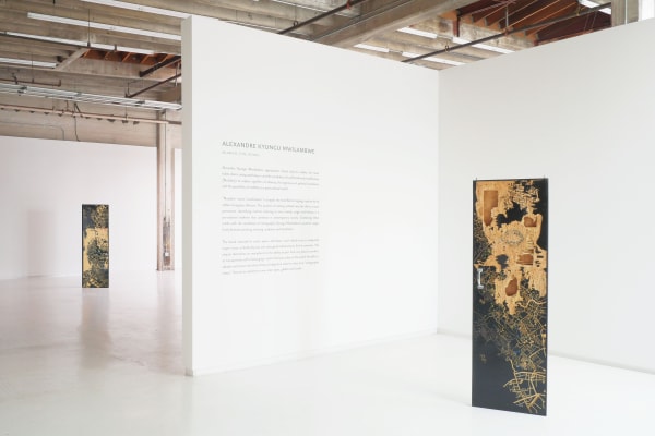 The image shows a minimalist art gallery installation featuring the works of Alexandre Kyungu Mwilambwe, with two intricately carved door-like sculptures displayed in a spacious, industrial-style setting. The artwork, titled "Mlango (The Door)," explores