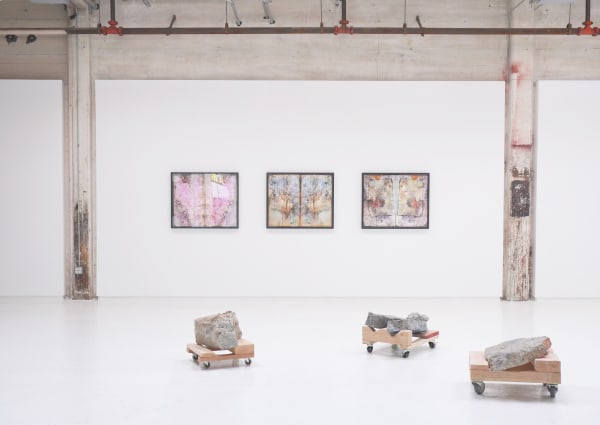 The image shows a contemporary art gallery installation featuring three framed abstract artworks hung on a white wall and three sculptural pieces on the floor. The artworks on the wall have a weathered, textured appearance with soft, muted colors and intr
