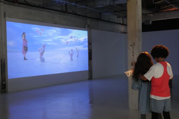 A sculpture of two girls holding a laundry basket faces a projected screen showing a girl walking through a salt field with blue sky and alien plants