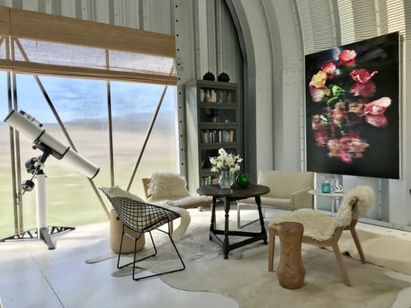 Rounded metal structure with telescope, hide rug and wooden living room set with plush pillows and large painting of pink petals by Cornelius Völker.