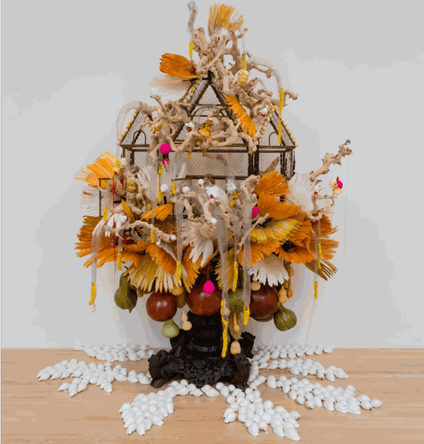 Sculpture with small white shells fanning away from small ornate carved wooden base adorned with yellow tail feathers, small metal birdcage and bits of wood.