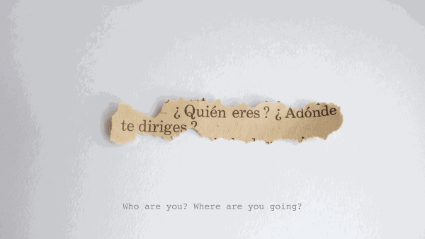 ripped scrap of brown paper with black text on white surface reads "¿Quién eres? ¿Adónde te diriges?" with caption underneath that reads "Who are you? Where are you going?"