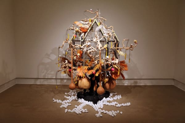 Rina Banerjee, Her captivity was once someone’s treasure and even pleasure but she blew and flew away took root which grew, we knew this was like no other feather, a third kind of bird that perched on vine intertwined was neither native nor her queens daughters, a peculiar other., 2011. Anglo-Indian pedestal (1860), Victorian birdcage, shells, feathers, gourds, grape wine, coral fractured charlotte doll heads, steel knitted mesh with glass beads, Kenyan tourist sculptures, and apple gourds. 84 × 84 × 72 inches. Courtesy of the San José Museum of Art.