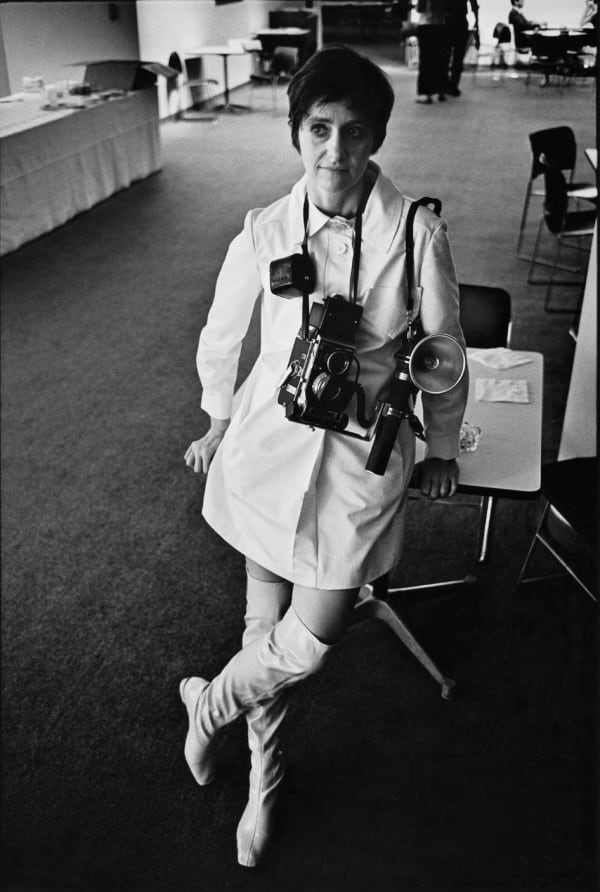 Diane Arbus with camera around her neck posing in white dress and white boots