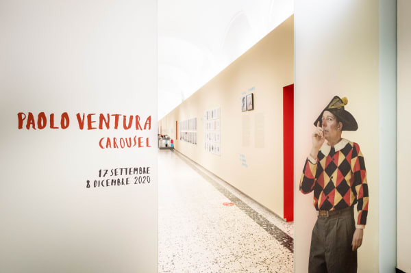 Paolo Ventura Carousel at CAMERA Center for Photography with hand-painted title and self-portrait on glass door with hanging artworks in yellow exhibition hall