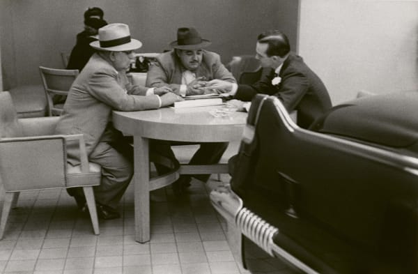 three white men in suits and hats sitting around a round table in a Cadillac showroom with the rear fin of a Cadillac in the foreground as if the fourth person at the table, by Robert Frank