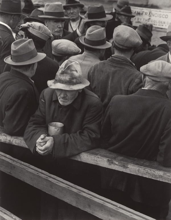 A black-and-white photograph by Dorothea Lange showing a group of men wearing hats and coats tightly gathered together, likely during the Great Depression. One elderly man stands at the front, leaning on a wooden railing with a weary expression, holding a