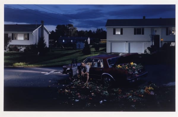 An image from Gregory Crewdson’s Dream House series depicting a nighttime suburban street. In the foreground, a man sits beside the open door of a maroon car, while the car’s trunk and the surrounding pavement overflow with scattered, colorful flowers. Tw