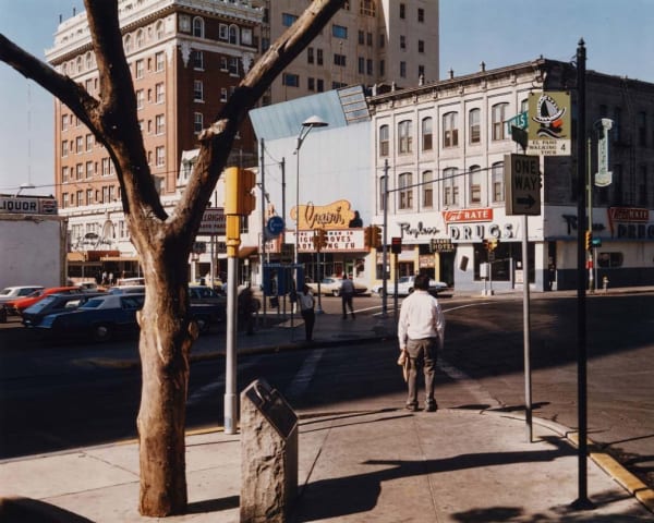 Man stands at a sunlit street corner in vintage downtown El Paso in this Stephen Shore photograph featuring cars and old storefronts.