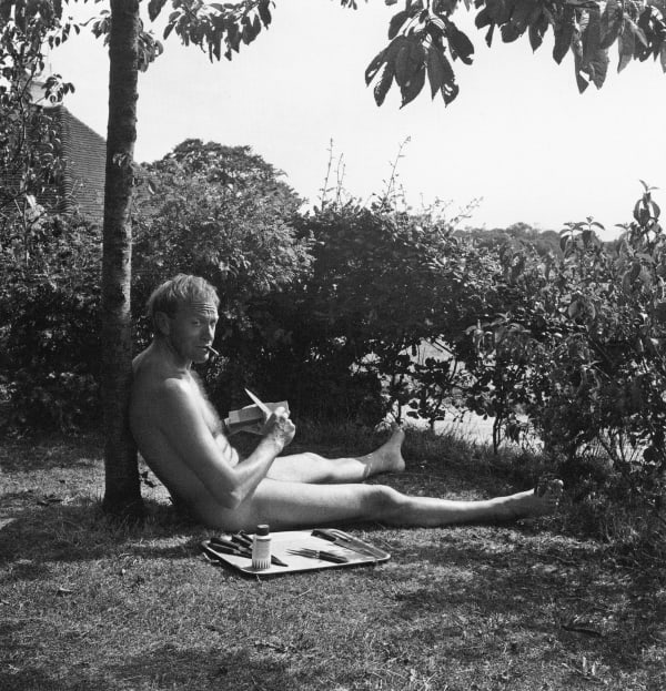 Black-and-white photograph of sculptor Lynn Chadwick sitting naked against a tree at Farley Farm, East Sussex, in 1957, sharpening kitchen knives laid out on a tray before him. Photograph by Lee Miller © Lee Miller Archives.