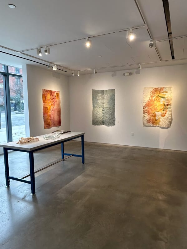 photo of an installation with three large works on paper in front of a table. ON the table there are multiple small sculptures laid out. There are windows in the left of the image, letting in natural light.