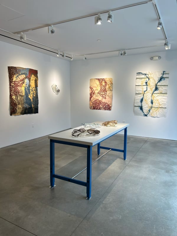 photo of an installation with three large works visible, as well as one sculpture on the wall with them. in the center of the room is a table, with several other sculptures laid out.