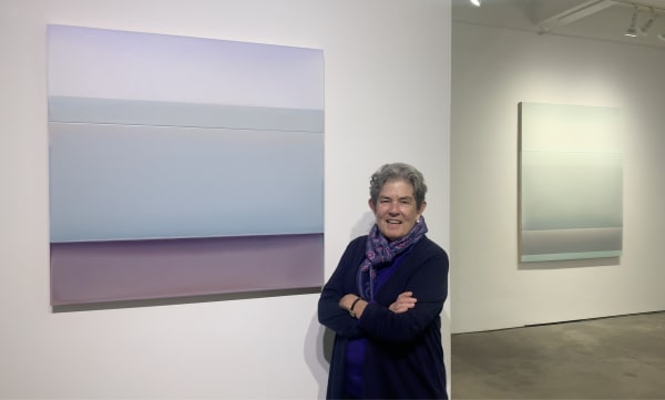 Woman standing in front of a pale purple and blue abstract painting hanging on a gallery wall.