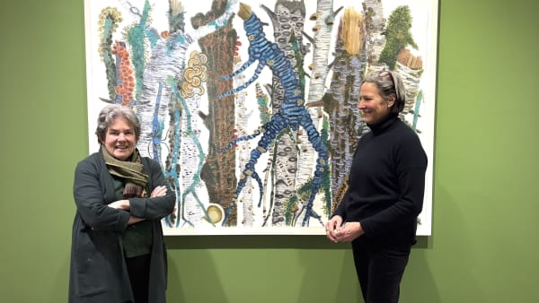 Two women standing in front of a large painting of colorful tree branches with fungus, mushrooms and moss