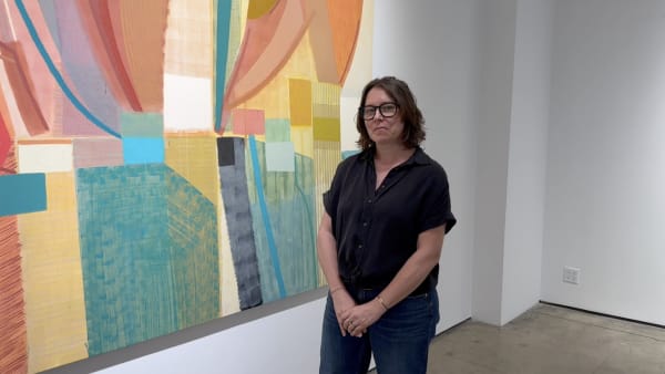 Woman in black shirt standing next to a large, colorful abstract art painting hanging on a gallery wall.