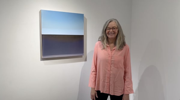 Female artist with gray hair in pink shirt standing in front of a blue abstract painting hanging on an art gallery wall.