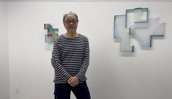 Male artist in striped shirt and glasses standing in front of gallery wall with two abstract wall sculptures