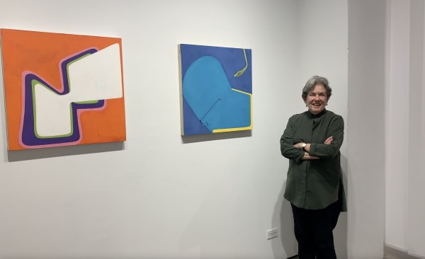 Woman standing next to two colorful, geometric, abstract paintings hanging on a gallery wall.