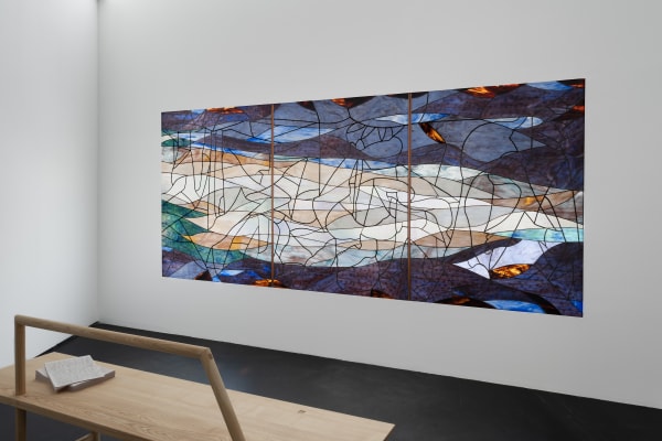 Installation view five | rindon johnson | Photo: Marjorie Brunet Plaza. Wall with a dark blue, green and orange stained glass window made up of 3 panels and a wooden bench with a stack of paper handouts on it.