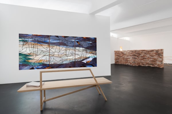 Installation view five | rindon johnson | Photo: Marjorie Brunet Plaza. White room with a dark blue, green and orange stained glass window made up of 3 panels on a plain wall, a red-brown brick wall behind it and a dark floor.