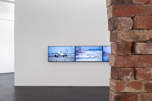 Installation view five | rindon johnson | Photo: Marjorie Brunet Plaza. White wall with three screens, streaming airport scenes, behind the side of a red-brown brick wall.