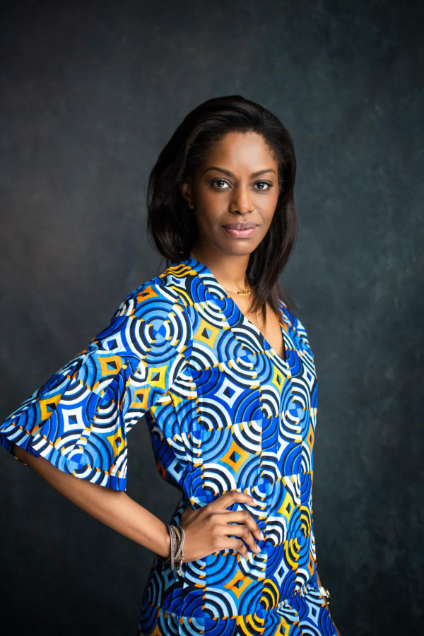 Portrait of a confident woman wearing a vibrant blue and yellow geometric-patterned, African-style, dress. She has a poised stance with one hand on her hip, showcasing a sophisticated style and an assertive demeanor against a dark textured backdrop.