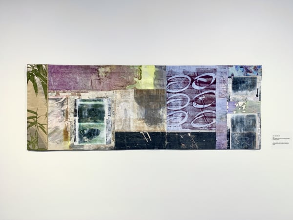 This quilt is a long rectangle, suggesting a scroll. It is made of many different sized, smaller rectangles overlapping and stitched as if collaged. The quilt’s predominant colors are gold and purple, with areas of black, white and grey. Several of the re