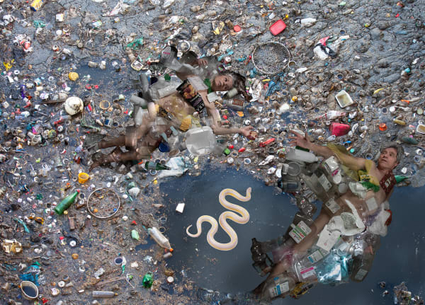 In this photograph, two nude figures, a man and a woman, lie on their backs on a trash-covered beach at low tide, each reaching a hand out to the other. Plastic containers and packaging are piled on, and embedded in, the sand that surrounds the figures, f