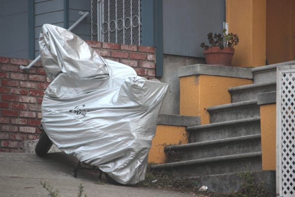 A photograph of a hilly city street, on which a parked scooter stands at the foot of a concrete stoop and in front of a brick wall. The scooter is fully covered in a silver plastic cover marked with the cursive Prima logo. The short walls of the stoop are