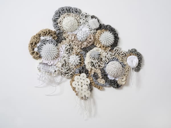 This work is made of crocheted wavy, curled, and coiled strips of white, off-white, beige, and gray yarn pinned on edge to the wall with pieces closely touching. The crochet surrounds two styrofoam half spheres covered with coils of yarn held flat to the