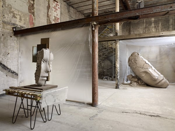 image of Mark Manders installation with split clay head on table and large dry clay head with plastic