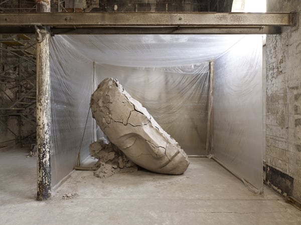 image of Mark Manders installation with split clay head on table and large dry clay head with plastic
