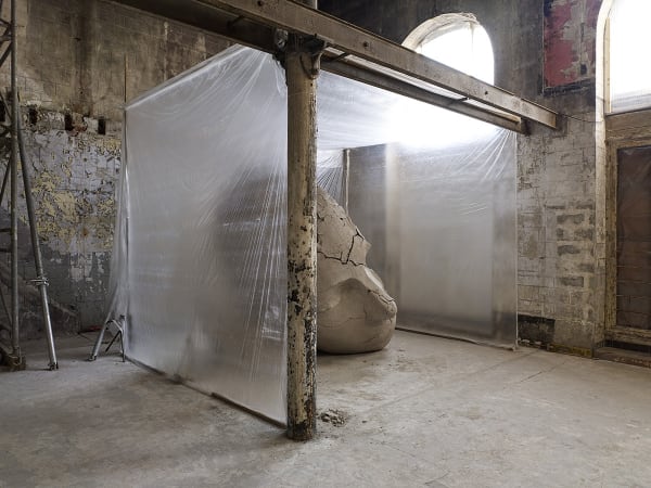 image of Mark Manders installation with split clay head on table and large dry clay head with plastic