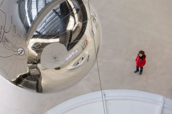 Person in a red coat looking up at the giant silver sphere