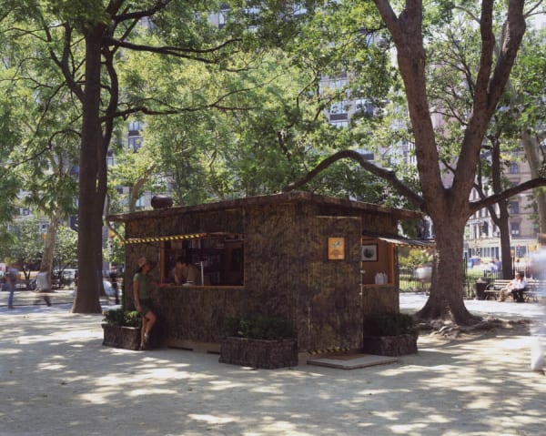 Installation image of the artwork in the park. A square camouflage stand under trees. A person leans out a window to talk to a person wearing shorts learning against the roofed structure.