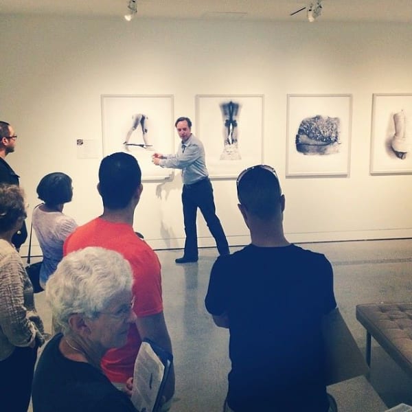 A man gestures to a painting while a crowd observes him.
