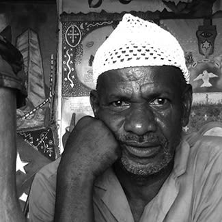 Black and white photo of a man looking at the camera. He has a white skull-cap and his chin is rested on his fist.