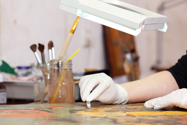A person in white gloves working under a light to restore an artwork alongside a glass jar of tools and brushes.