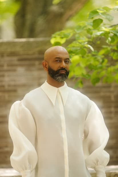 Headshot of Brendan Fernandes in white, standing in front of a stone wall and tree
