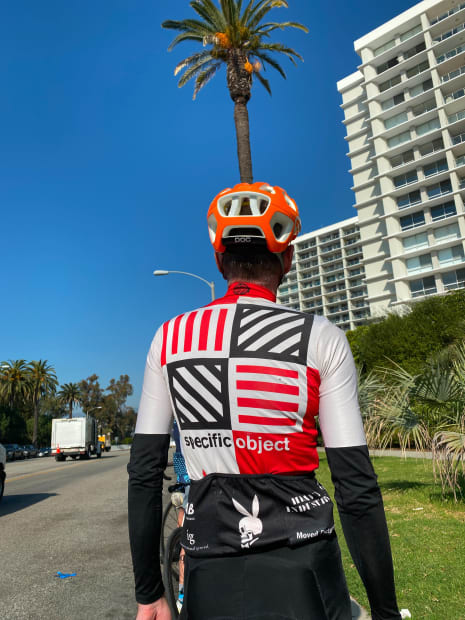 Behind of a man wearing a bicycle helmet and jersey, palm tree in the background
