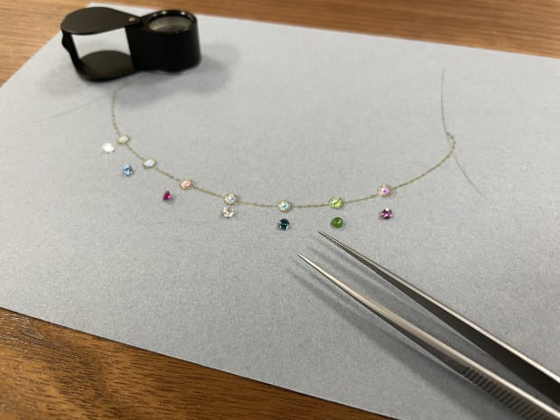 Loose rare gemstones alongside a hand-painted rendering of an acrostic necklace and jeweller’s loupe, showcasing bespoke jewellery design process.