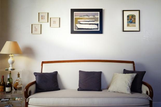 An elegant white sofa with curved wooden trim and blue patterned throw pillows, positioned beneath a gallery wall of framed artwork and adjacent to a modern glass bar cart with a lamp and bottles.