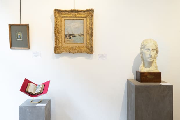 Gallery installation view at La Gabrielle Fine Arts: maritime painting in ornate gilt frame, illuminated manuscript on red stand, and plaster bust on concrete pedestal against white wall.
