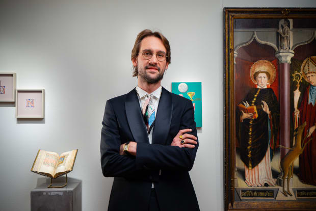 Professional art gallery interior featuring an elegantly dressed gallery director in a dark suit and patterned tie, standing with crossed arms against a white wall. The space showcases medieval art pieces including an illuminated manuscript on a display s