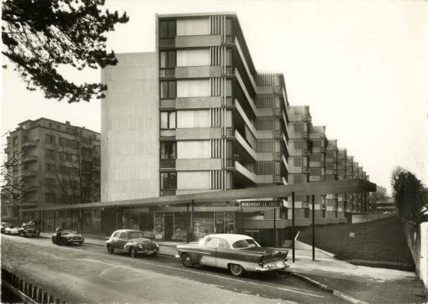 Vintage black and white photograph of a mid-century modern apartment building with classic cars parked in front.