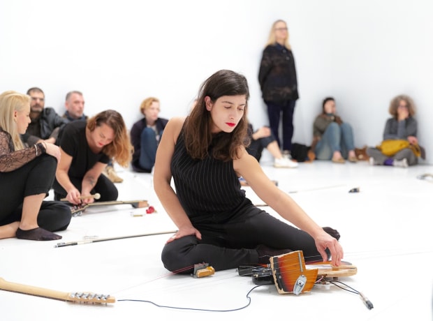 Photo of Naama Tsabar performing. She's sitting on the floor strumming guitar