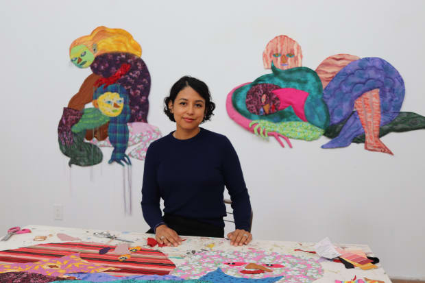 Portrait of Maria A. Guzmán Capron sitting at her table with two wall sculptures behind her