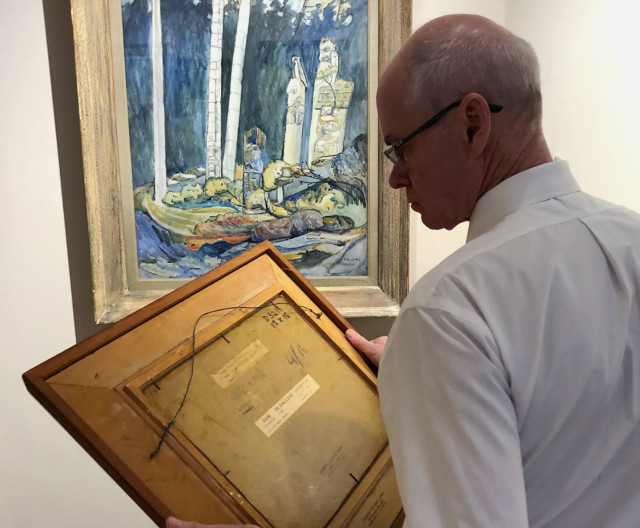 Alan Klinkhoff examining the back of a Lawren Harris painting with an Emily Carr watercolour hanging in the background.