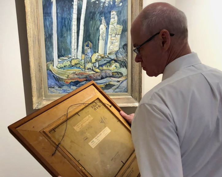 Alan Klinkhoff examining the back of a Lawren Harris painting with an Emily Carr watercolour hanging in the background.